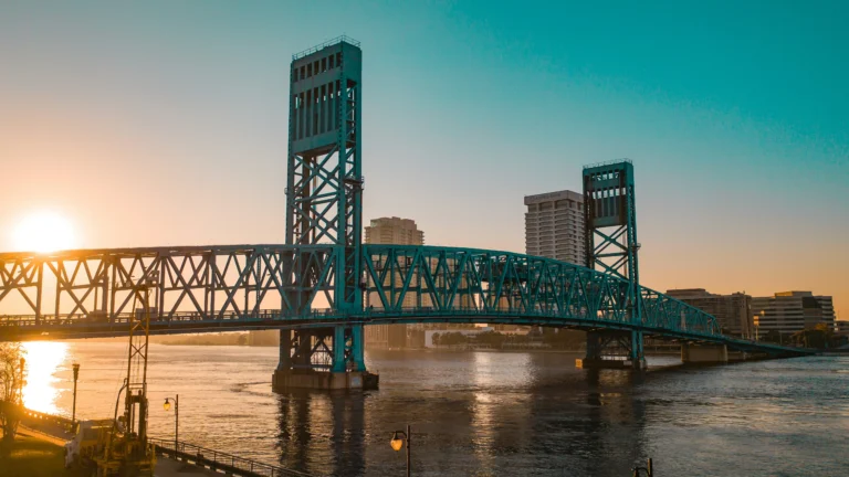 A beautiful sunset view of the iconic blue Main Street Bridge (John T. Alsop Jr. Bridge) stretching across the St. Johns River in downtown Jacksonville, Florida, with the bright sun low on the horizon casting a golden glow on the water and skyline.
